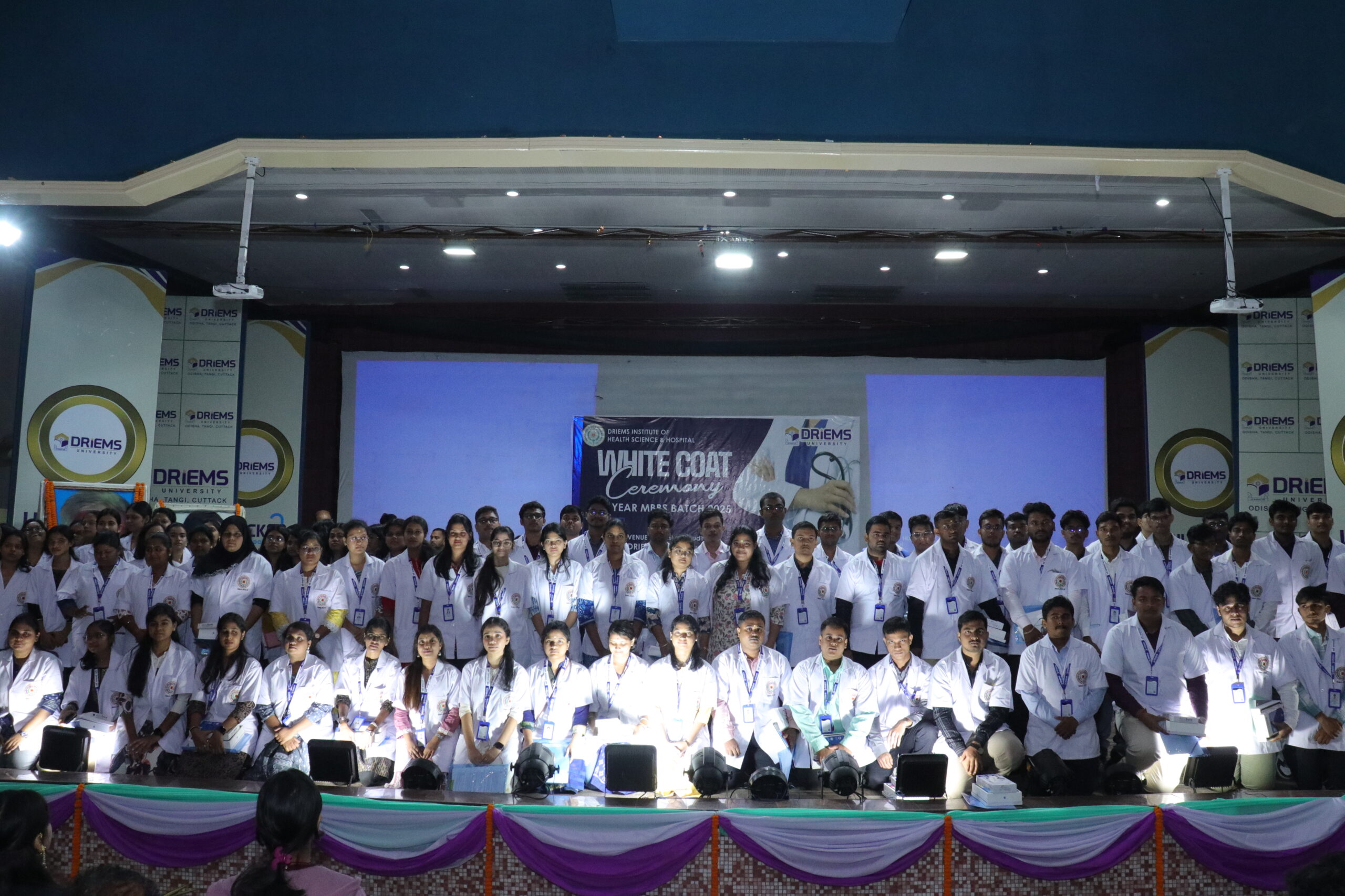 person writing in a notebook beside by an iPad, laptop, printed photos, spectacles, and a cup of coffee on a saucer